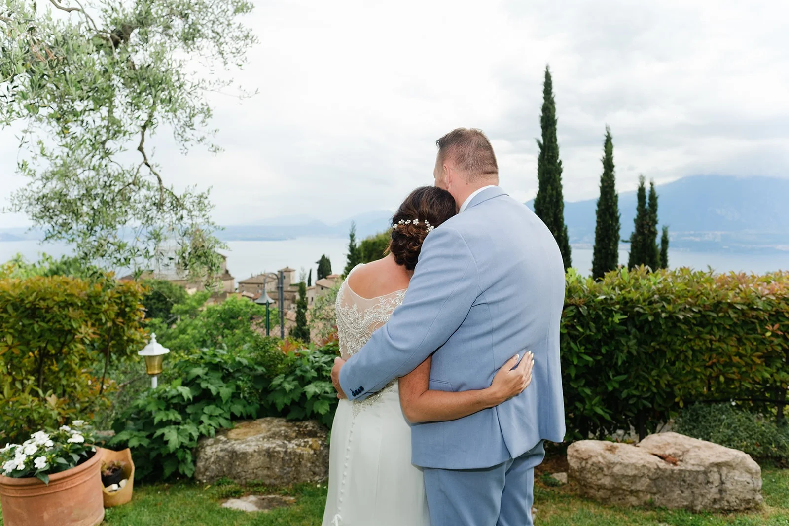 Sposi abbracciati di spalle, affacciati sul Lago di Garda fra gli ulivi